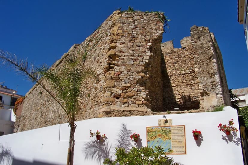 Muro de piedra del Castillo de San Luis visto desde la Calle Castillo, con panel informativo y jardín de acceso